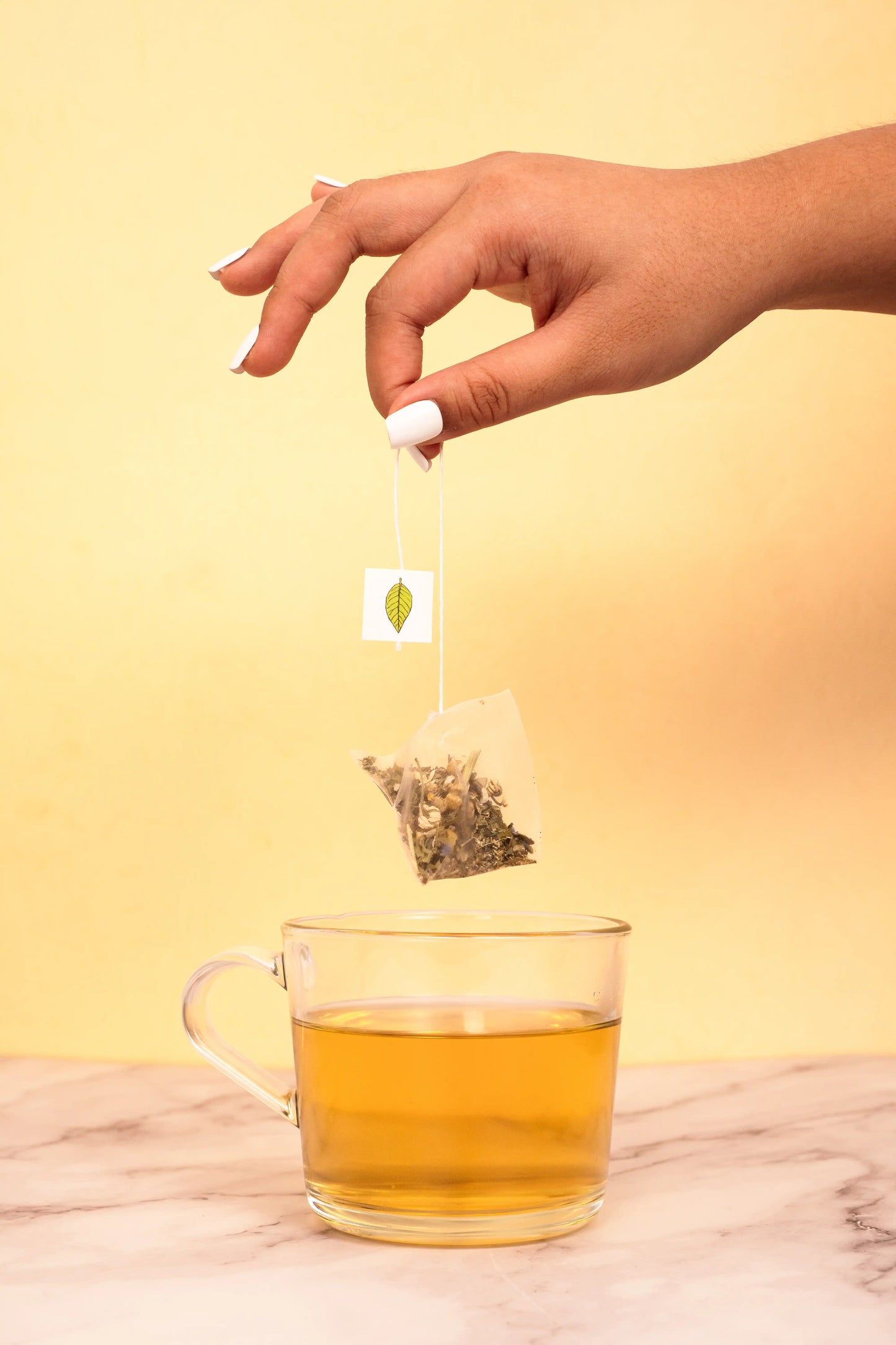 Hand placing a tea bag into a glass of tea on a yellow background