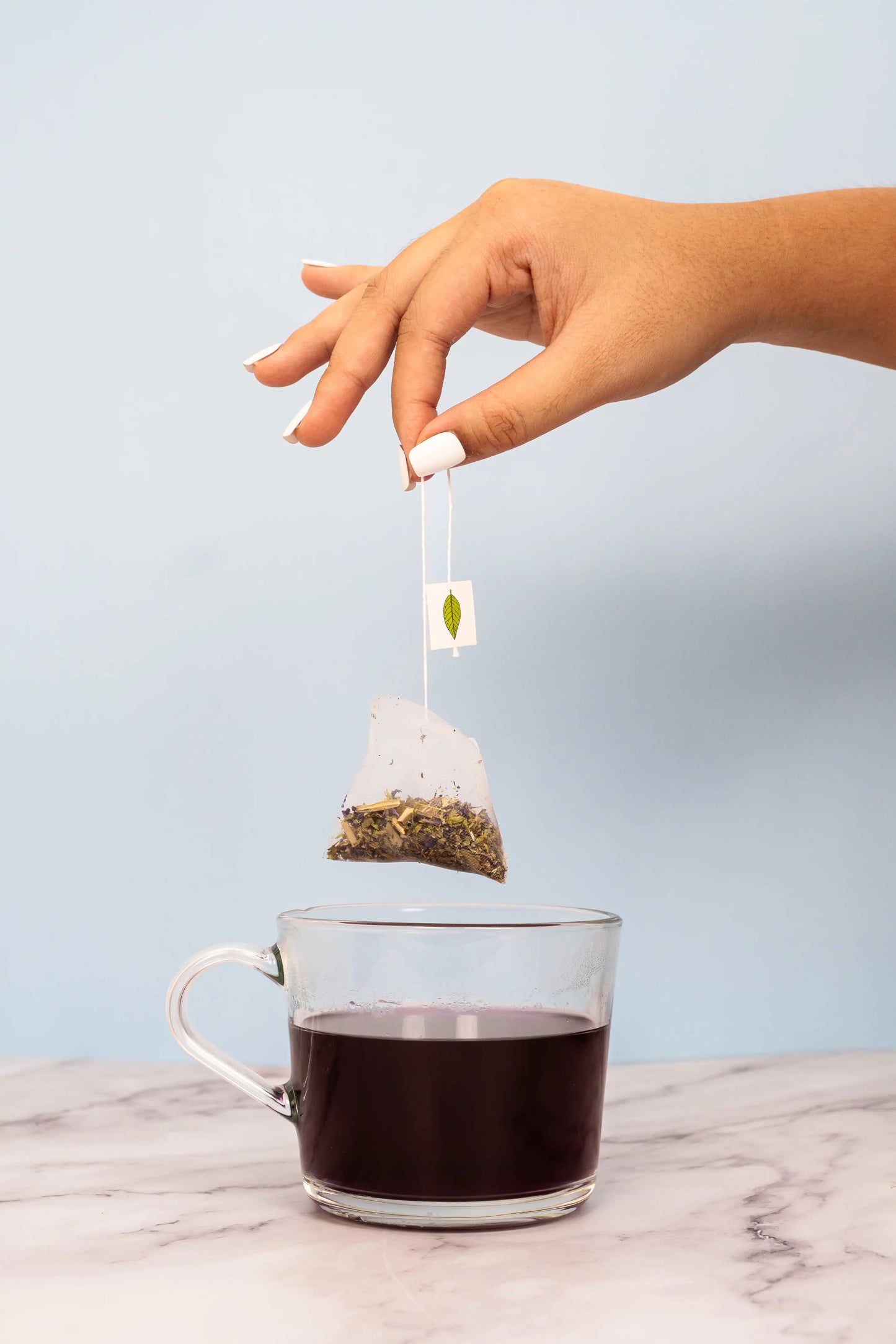 Hand placing a tea bag into a glass of black tea on a marble surface with a light blue background
