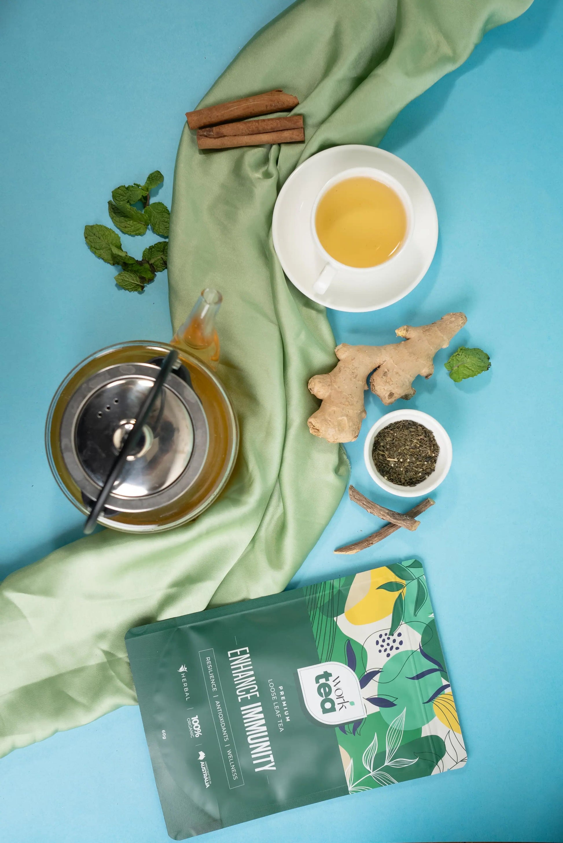 Tea-making setup with a green tea package, tea leaves, ginger, cinnamon, and a cup of tea on a blue background.